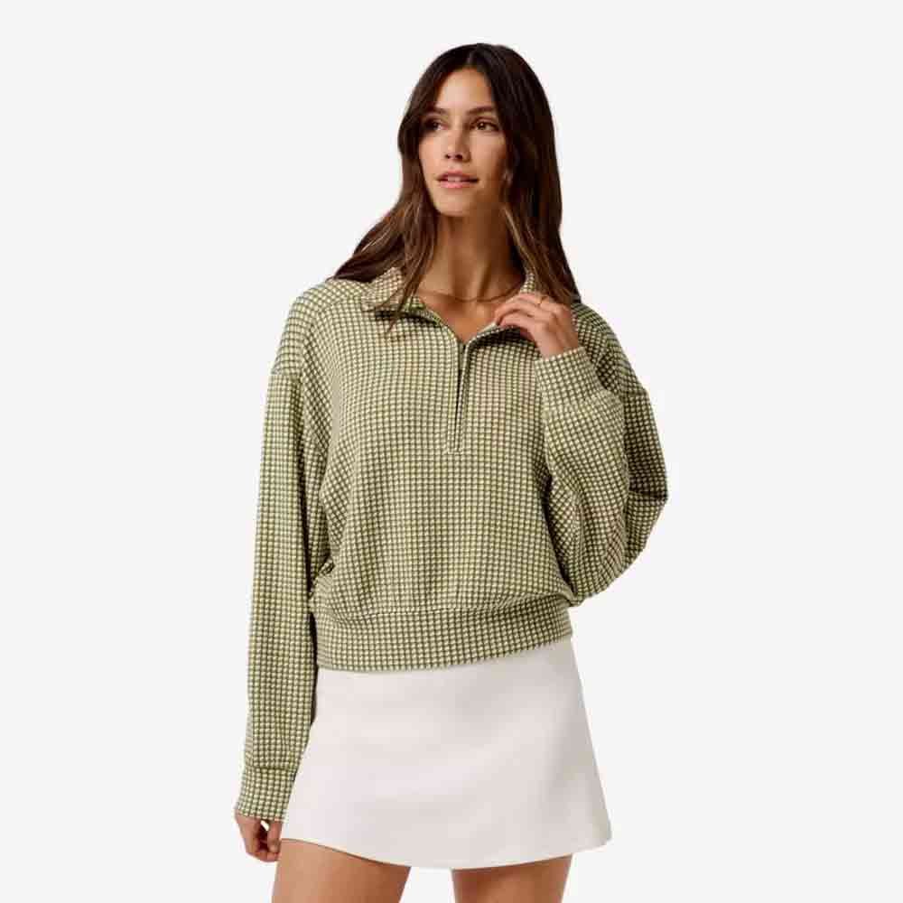 A woman wearing a green and white checkered long-sleeve pullover and a white skirt stands against a plain light background, looking slightly to the side—an elegant look perfect for Mother's Day.